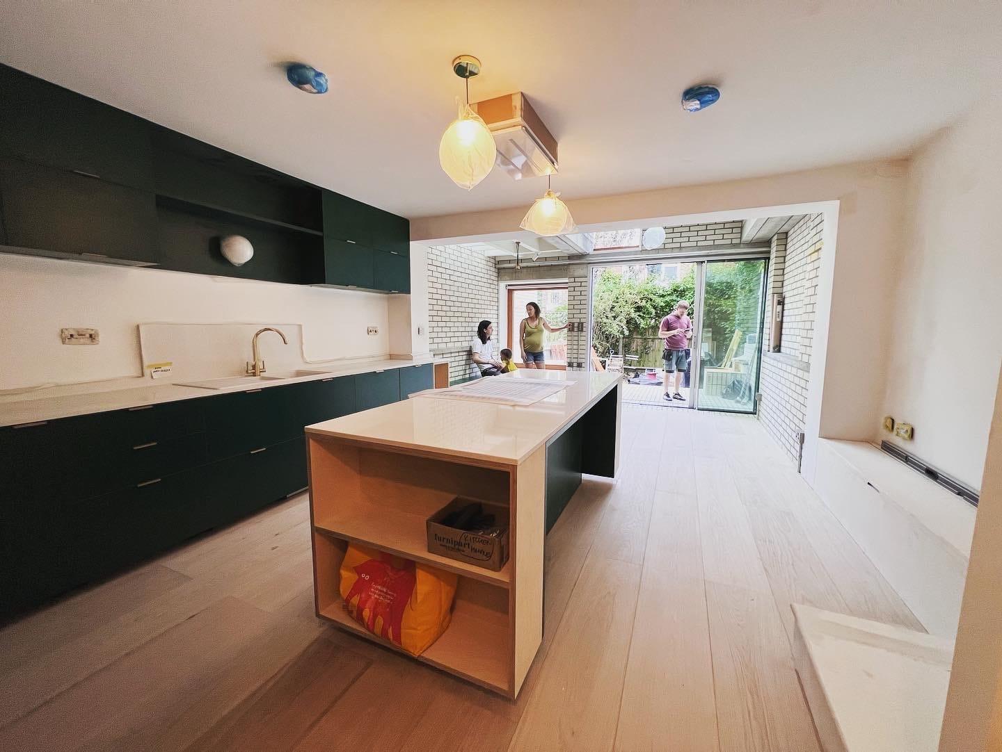 Dark-green kitchen with a birch-faced island, opening through full-width sliding doors onto the garden