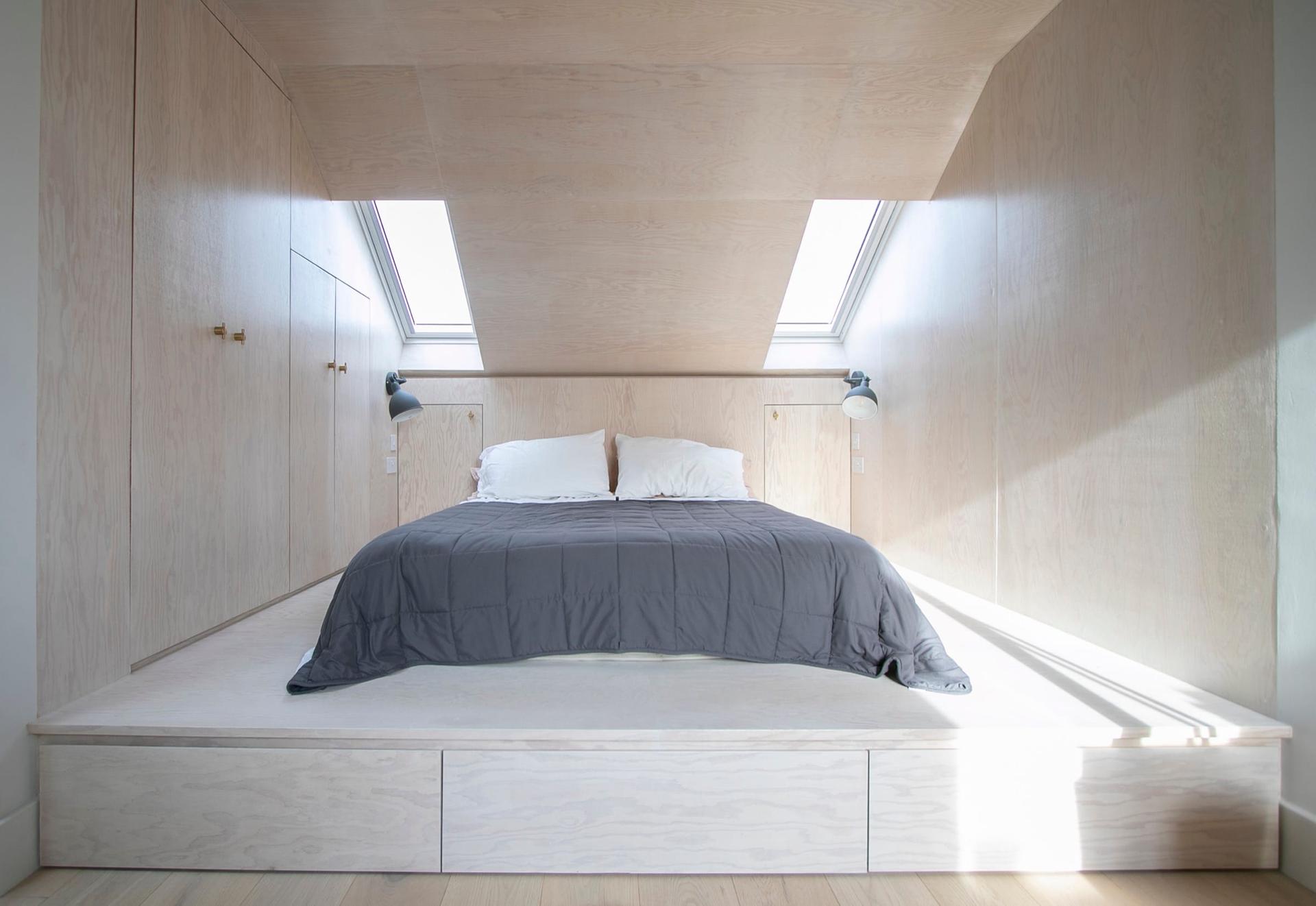 Loft bedroom lined in birch ply, the bed set on a raised platform with drawers beneath and rooflights above