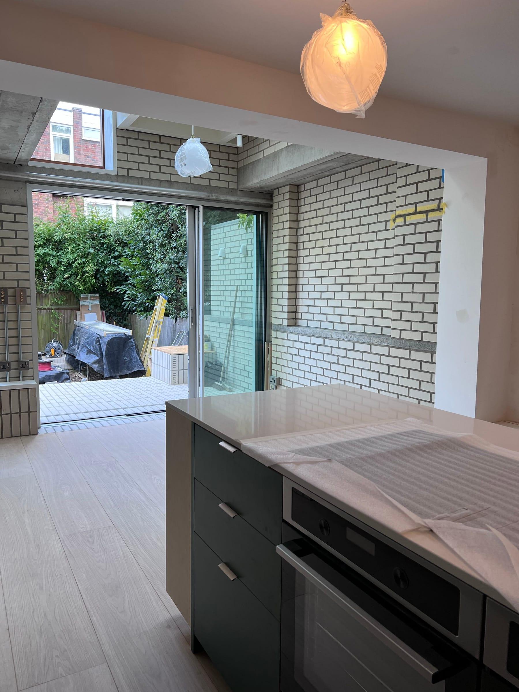 Kitchen island going in — worktop still filmed, dark units set, sliding doors open to the garden