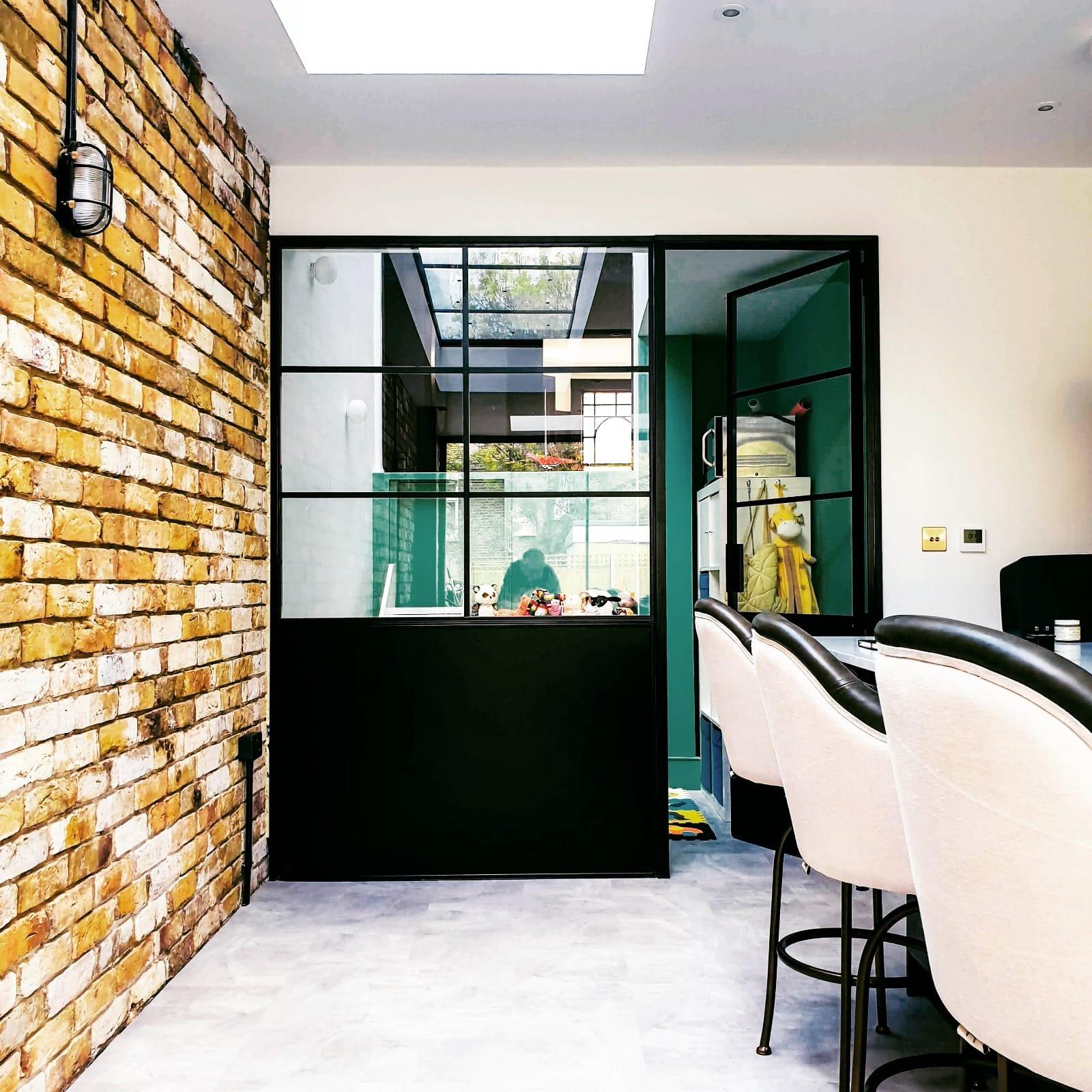 Kitchen against a yellow London stock brick wall, a black steel screen to a green room beyond and a rooflight over