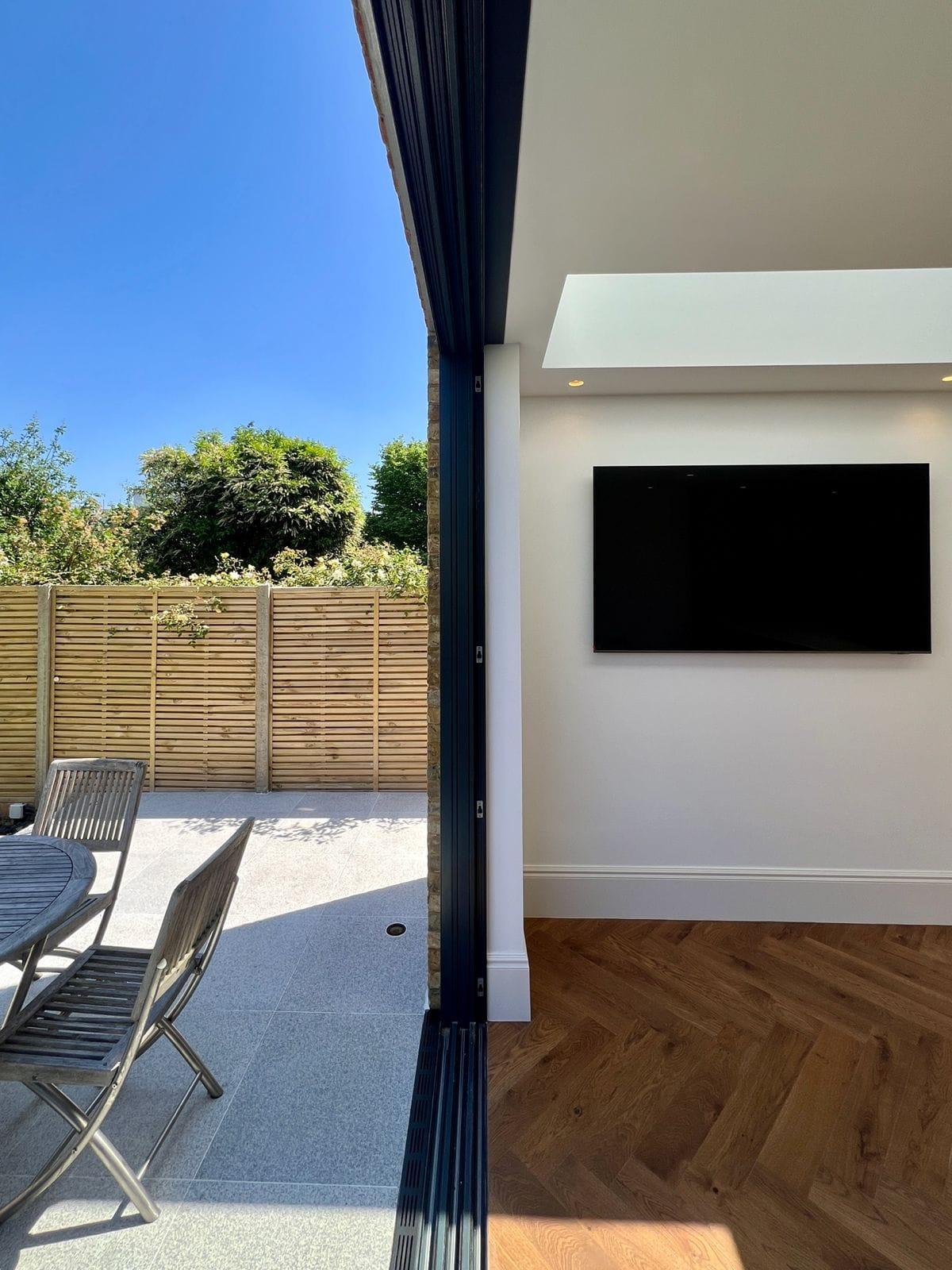 Bifold doors folded back onto a paved patio, with a wall-mounted screen and herringbone flooring inside