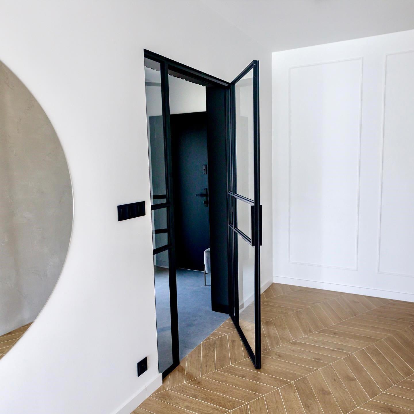 Black steel-framed door standing open onto a hallway, over chevron flooring