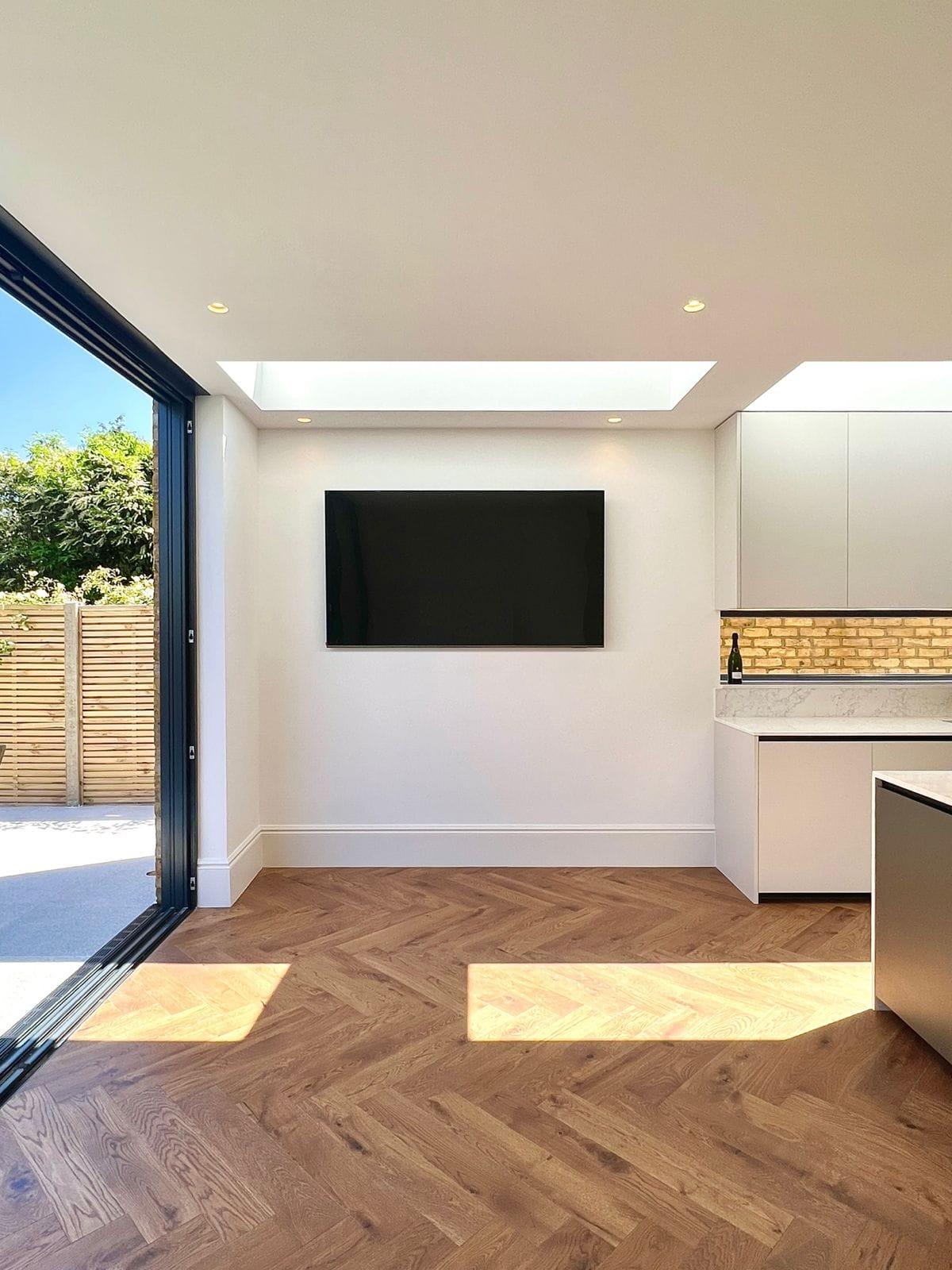 Rear extension interior with a white kitchen, a lit brick splashback and herringbone flooring to the garden doors