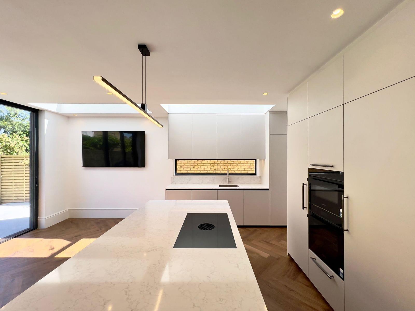 Kitchen island in a rear extension, with a lit brick splashback and integrated ovens in the run behind